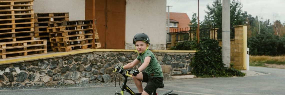 A young boy in a helmet rides his bicycle near a warehouse with stacked wooden pallets.