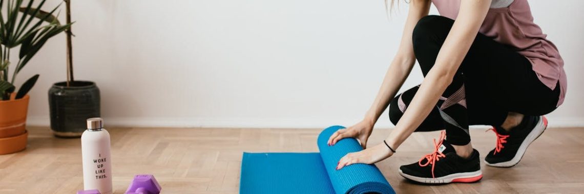 Side view of young female athlete in sportswear unfolding blue sport mat and looking down while sitting on floor near dumbbells and water bottle preparing for training indoors